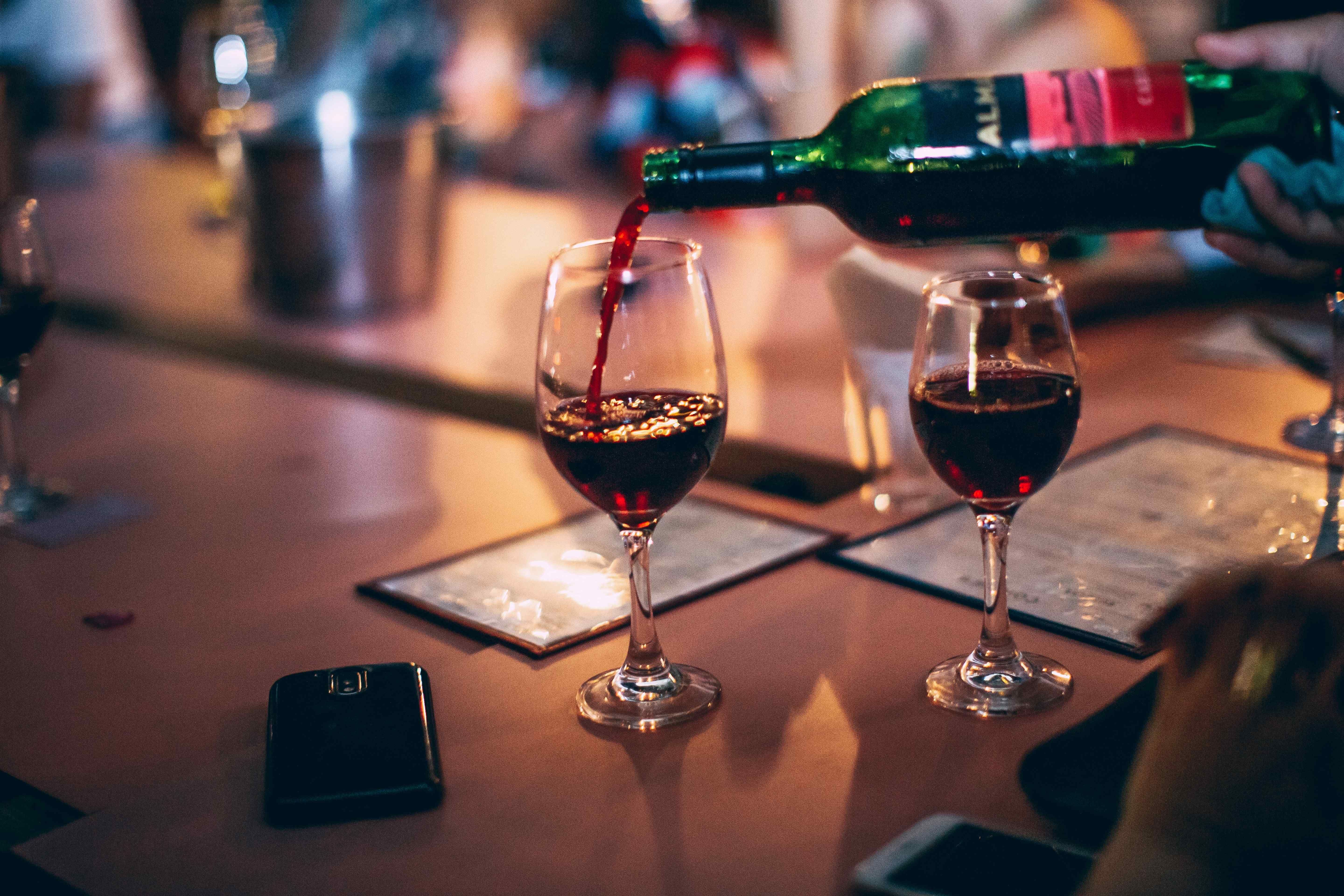 Red wine being poured into a glass at a bar, with a second full glass and phones resting on the counter.