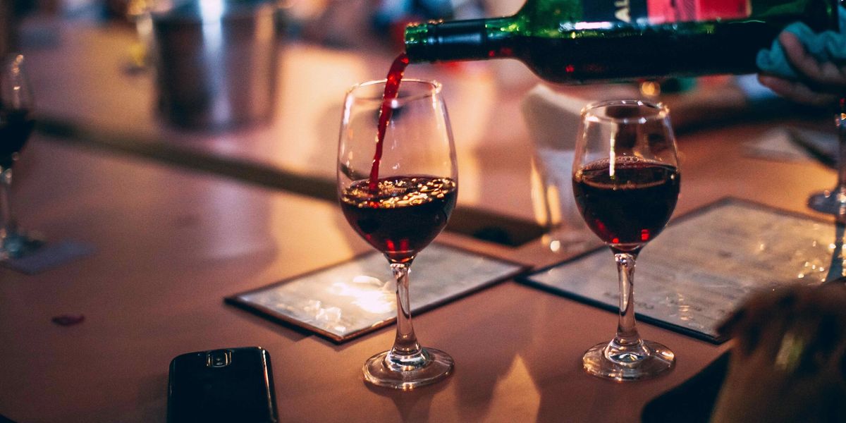 Red wine being poured into a glass at a bar, with a second full glass and phones resting on the counter.
