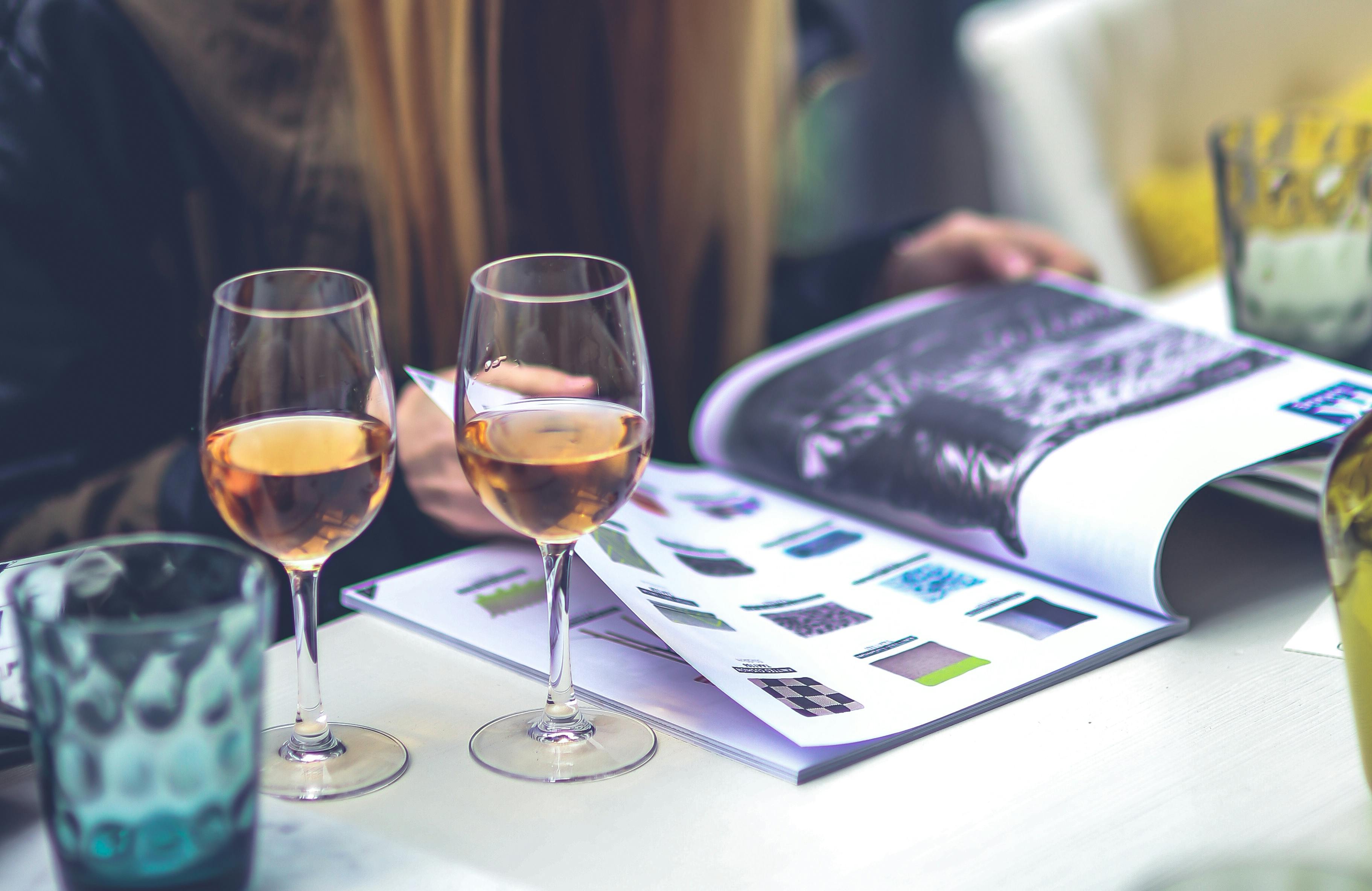A woman flips through a magazine as two glasses of orange wine rest on the table beside her.