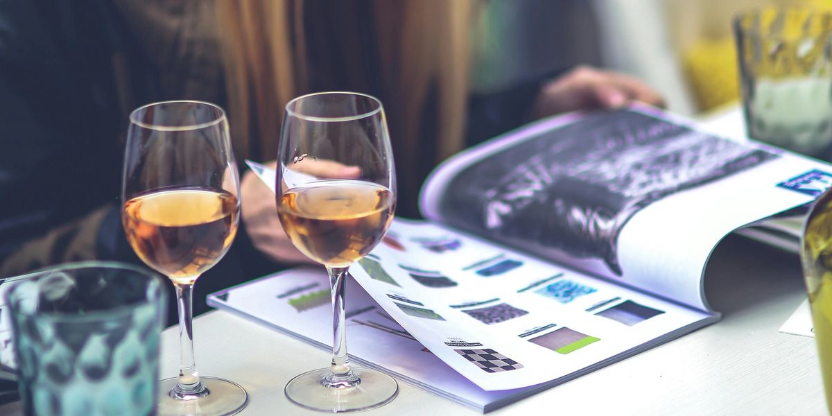 A woman flips through a magazine as two glasses of orange wine rest on the table beside her.