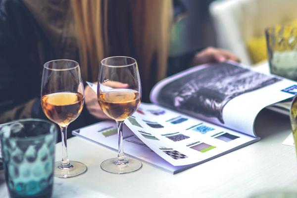 A woman flips through a magazine as two glasses of orange wine rest on the table beside her.