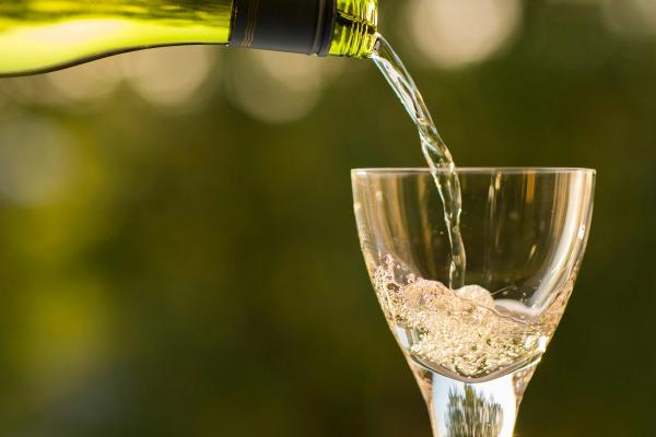 A wine glass being filled with white wine against the backdrop of a lush, green, outdoor environment.