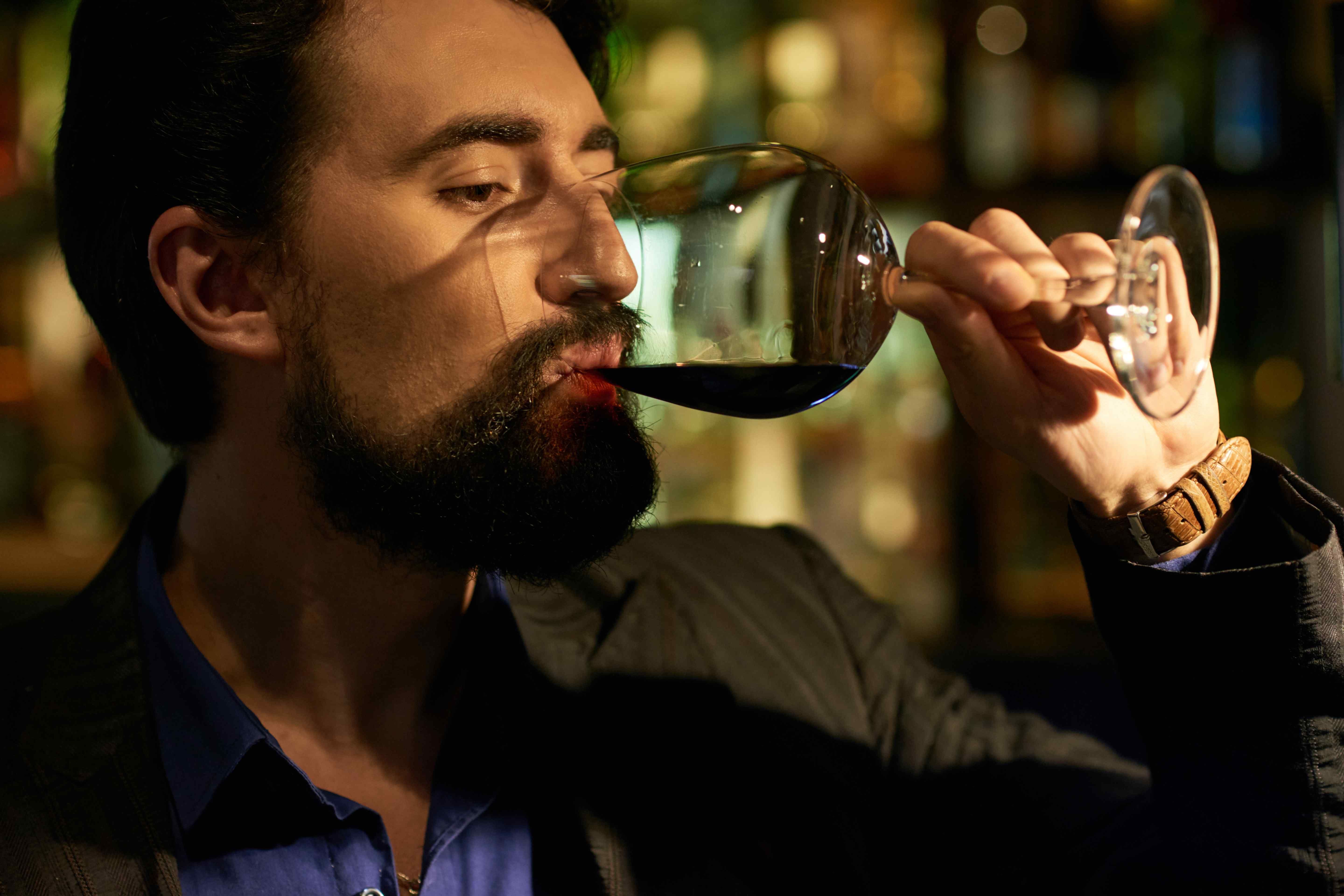 A bearded man in a dark jacket and blue shirt sips red wine from a large glass at a dimly lit bar.