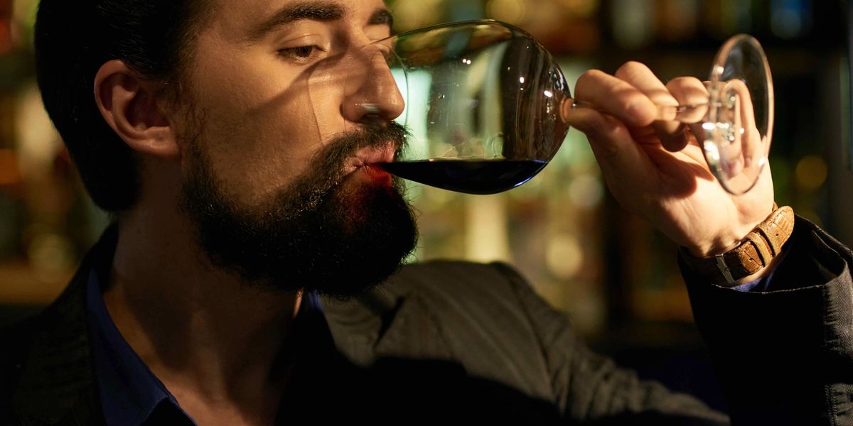 A bearded man in a dark jacket and blue shirt sips red wine from a large glass at a dimly lit bar.