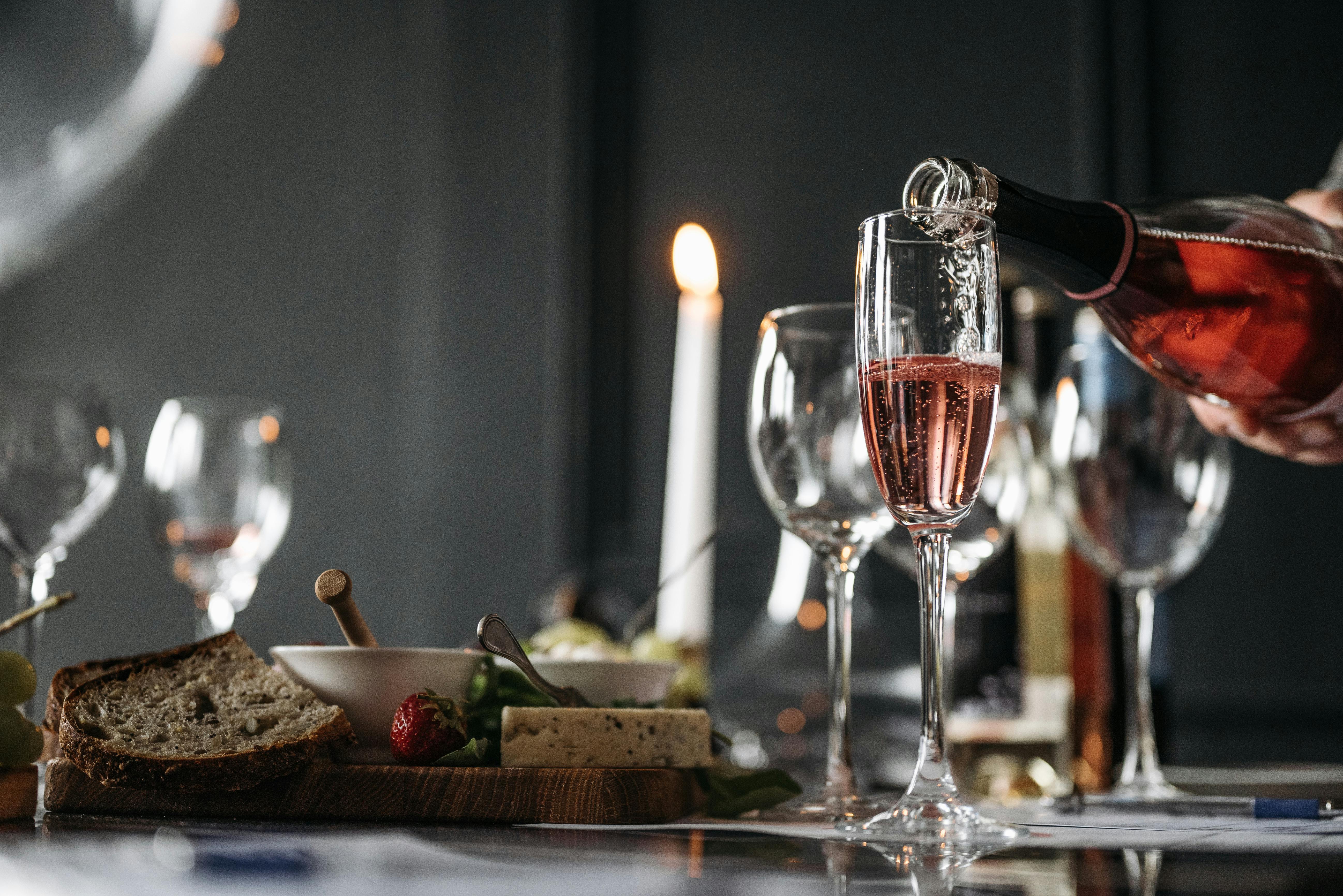 Sparkling rosé wine is poured into a Champagne flute on a table with bread, appetizers, and candles.