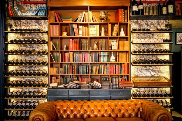 Open-shelf wine racks flanking a bookcase filled with books, decanters, and a leather sofa in a home library.