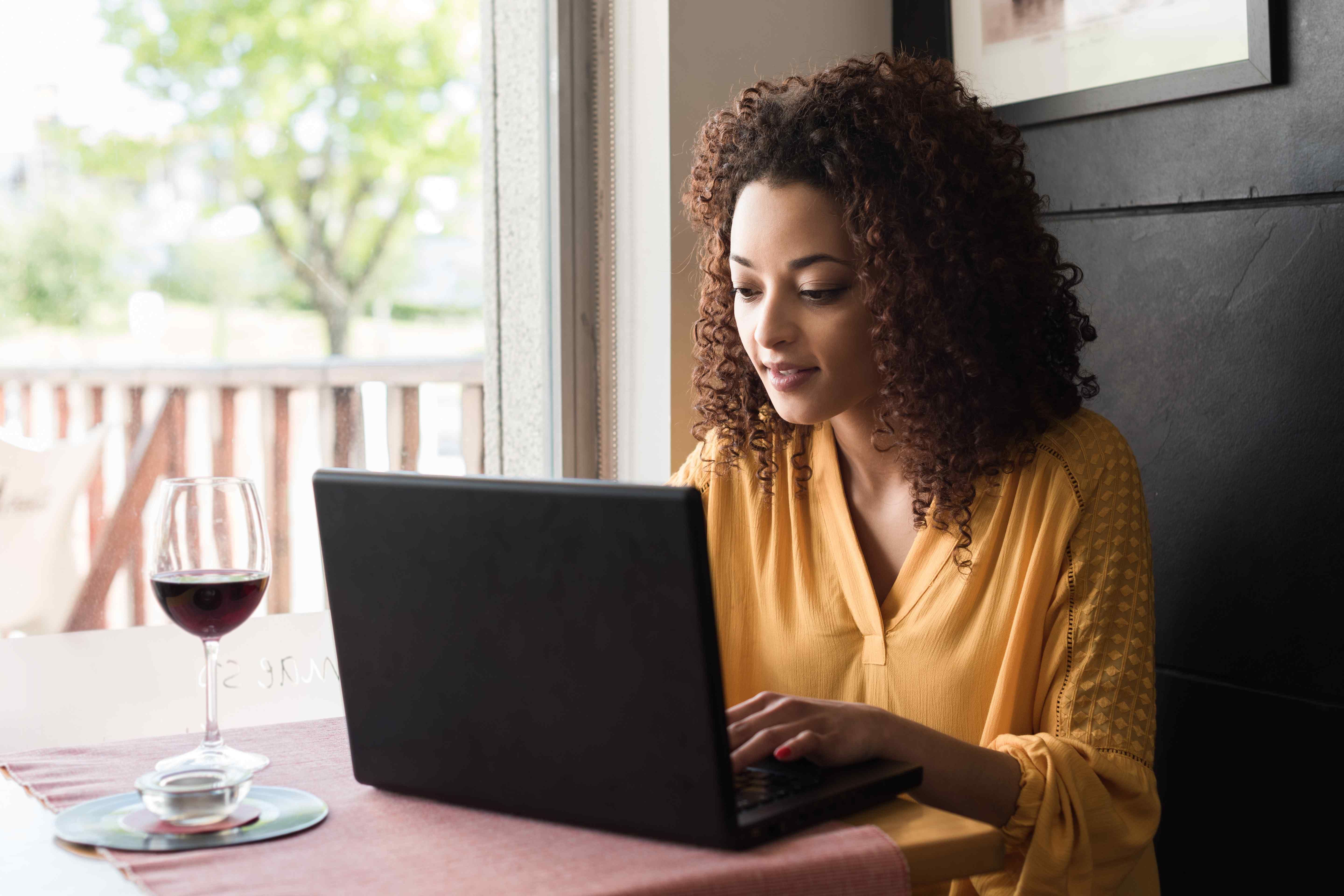 A woman using a wine collection app on her laptop with a glass of red wine beside her.