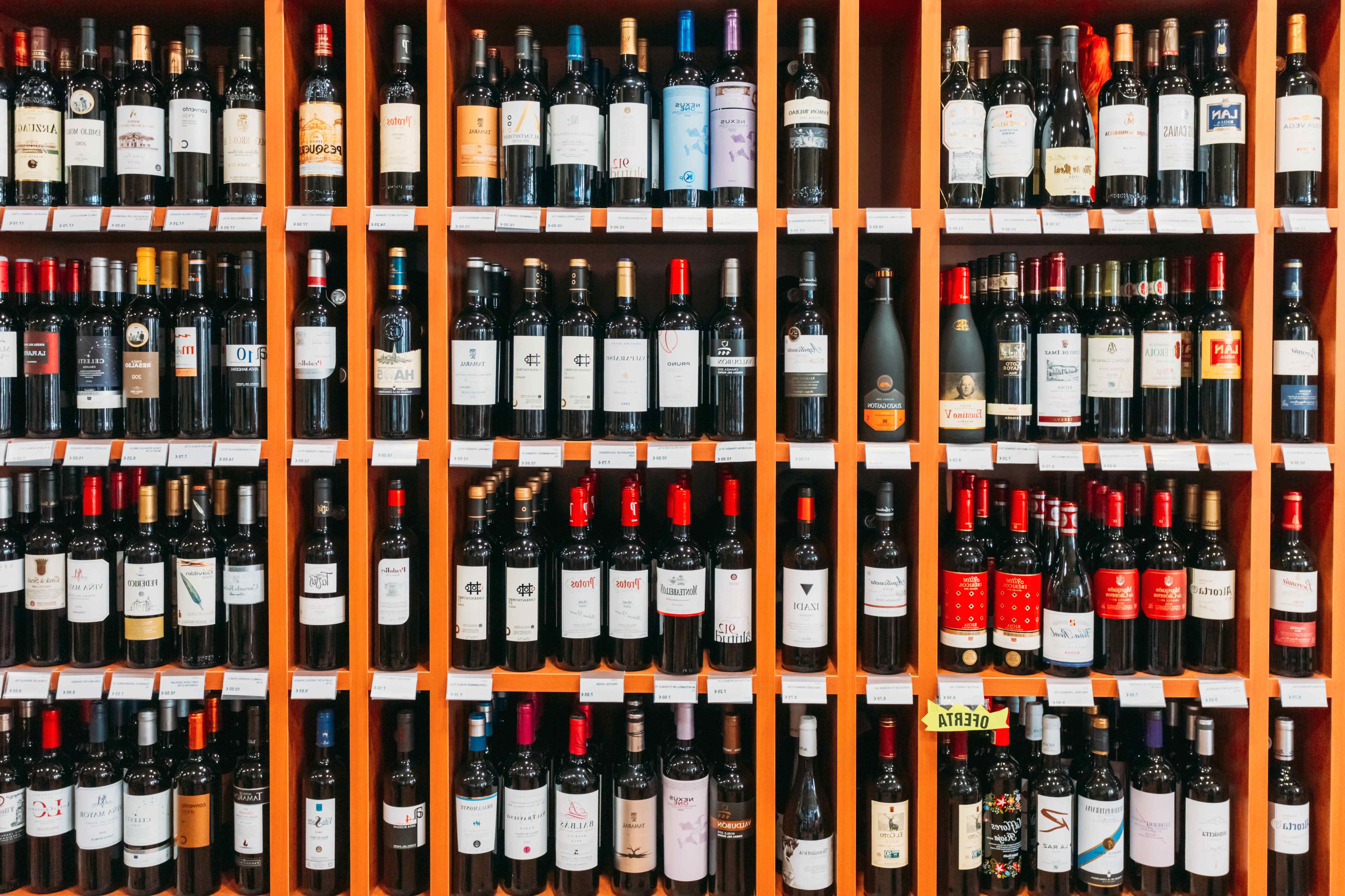 Rows of red wine bottles on orange wooden shelves in a wine shop, with small price labels along each shelf edge.