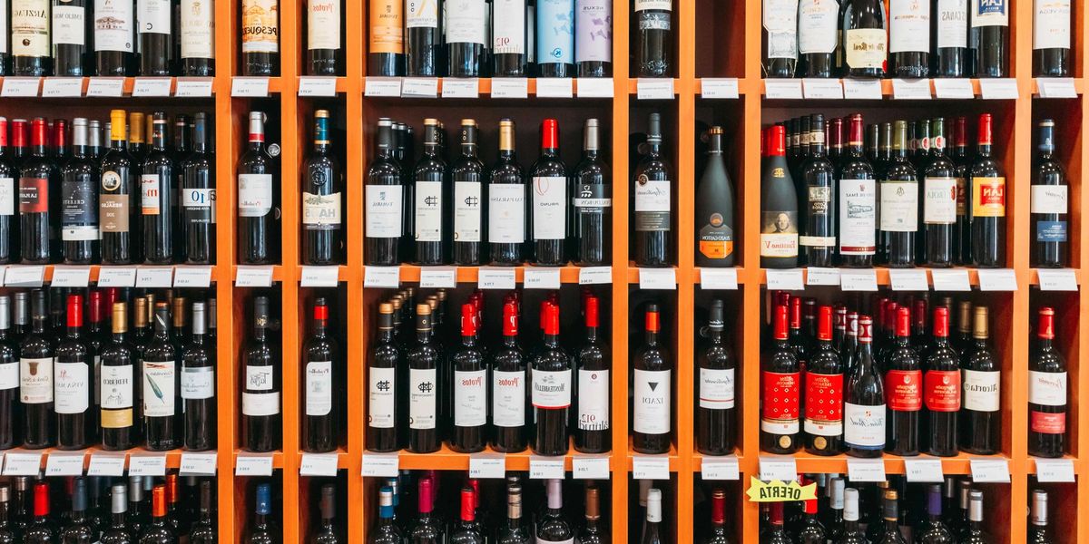Rows of red wine bottles on orange wooden shelves in a wine shop, with small price labels along each shelf edge.