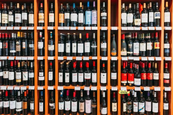 Rows of red wine bottles on orange wooden shelves in a wine shop, with small price labels along each shelf edge.