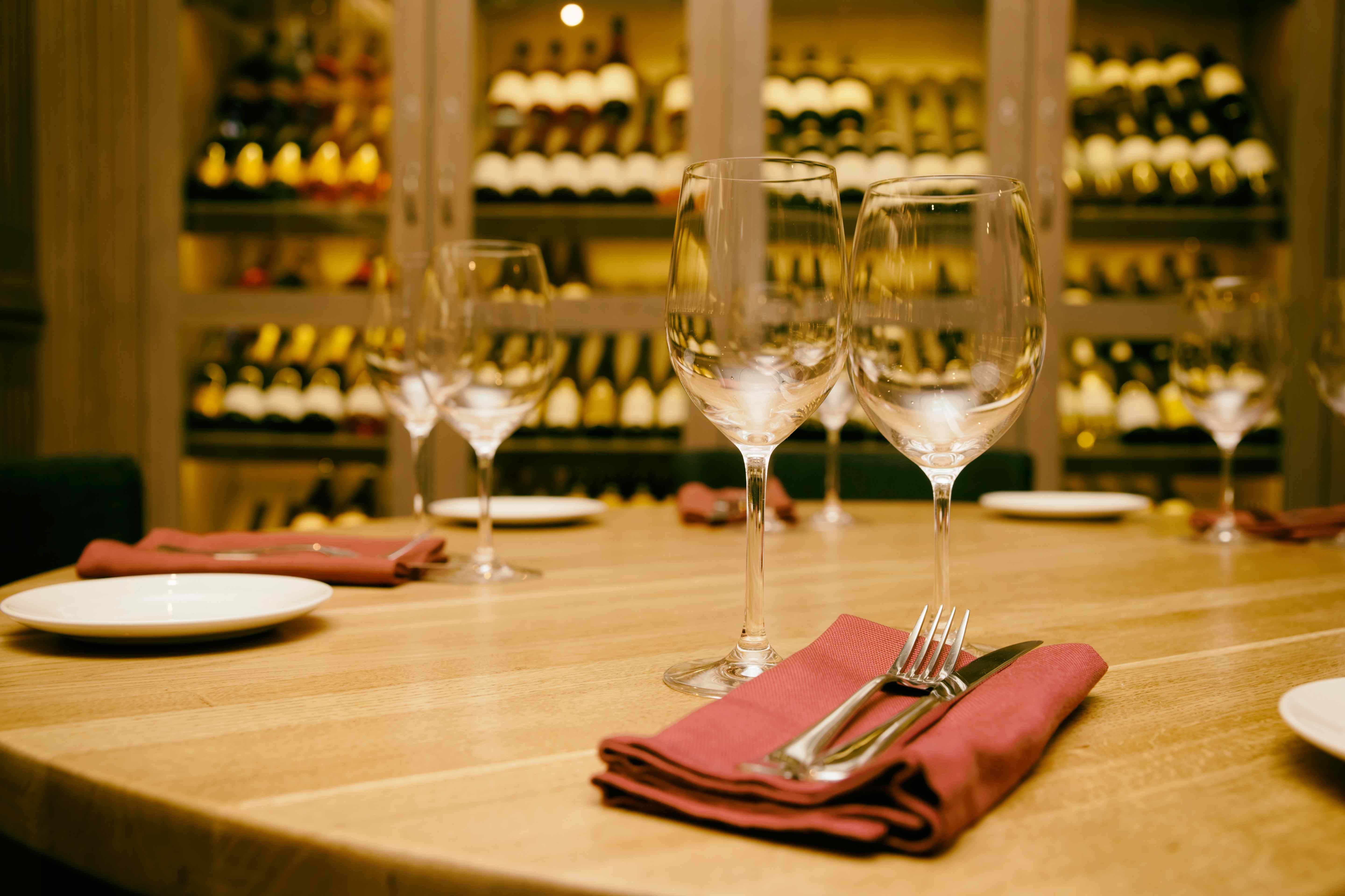 Elegant restaurant table set with wine glasses and napkins in front of a glass-enclosed wine cellar wall.