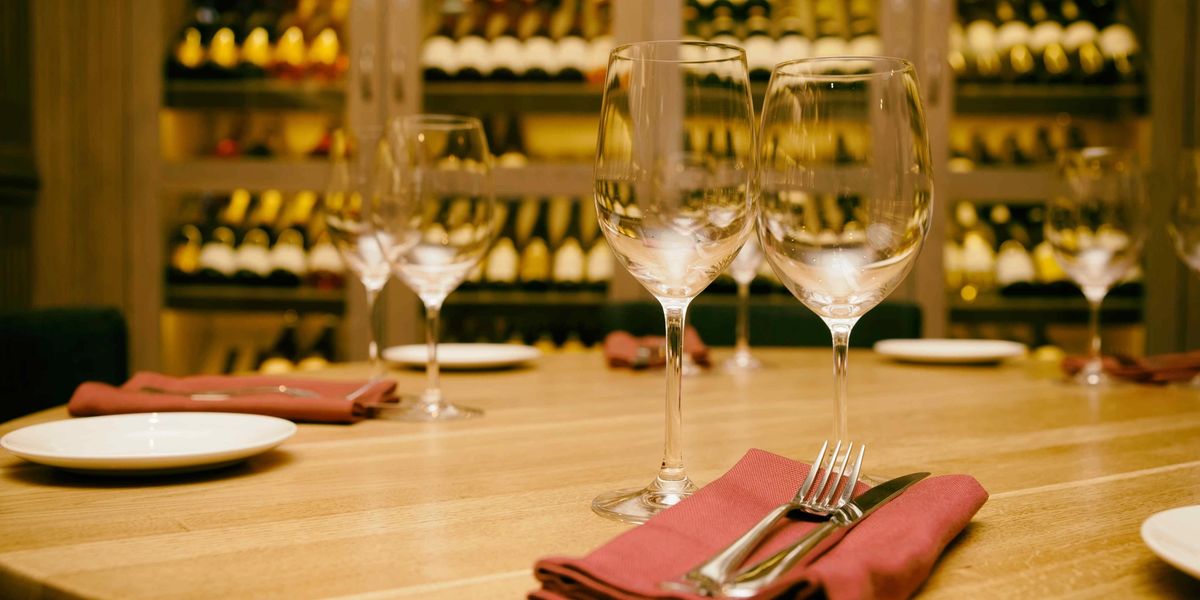 Elegant restaurant table set with wine glasses and napkins in front of a glass-enclosed wine cellar wall.