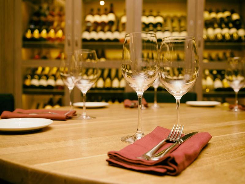 Elegant restaurant table set with wine glasses and napkins in front of a glass-enclosed wine cellar wall.