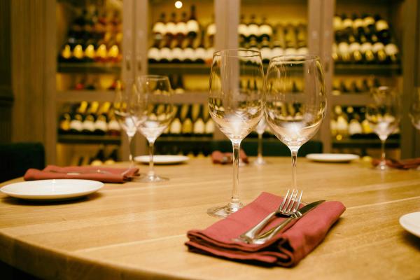 Elegant restaurant table set with wine glasses and napkins in front of a glass-enclosed wine cellar wall.