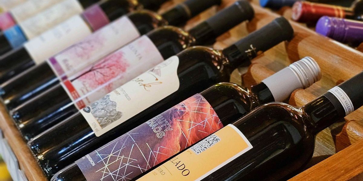 A row of red wine bottles, each with a colorful label, rest on a shelf in a wine shop.