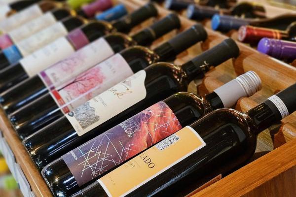 A row of red wine bottles, each with a colorful label, rest on a shelf in a wine shop.