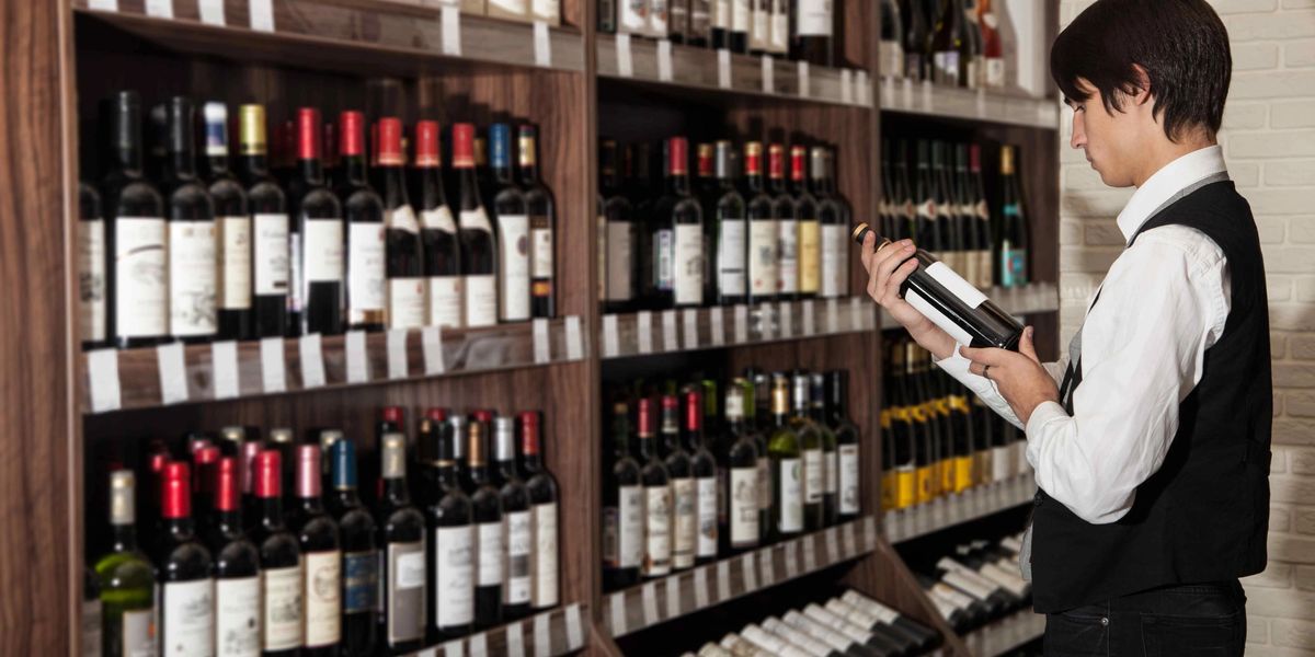 A sommelier in a vest examining a wine bottle's label while standing before fully stocked shelves.