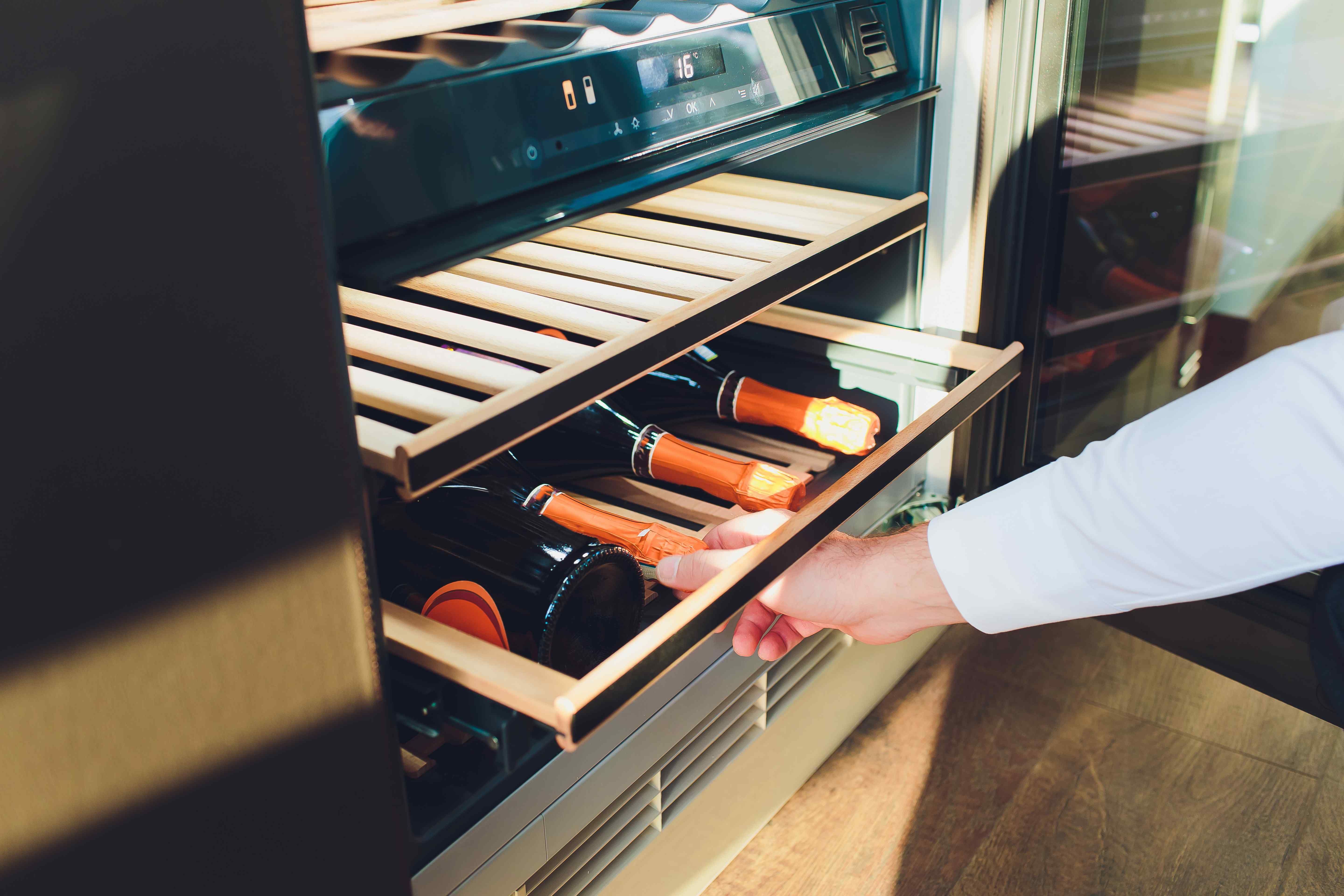 Hand pulling open a wine fridge drawer with a digital temperature display and champagne bottles inside.