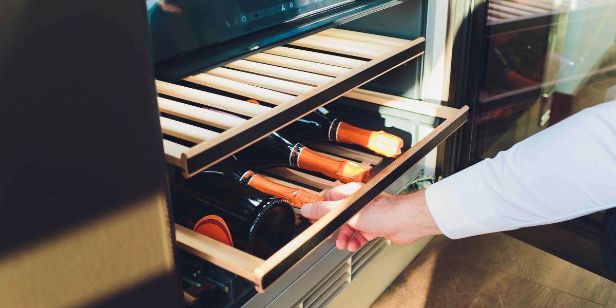 Hand pulling open a wine fridge drawer with a digital temperature display and champagne bottles inside.