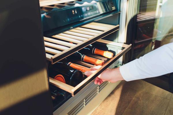 Hand pulling open a wine fridge drawer with a digital temperature display and champagne bottles inside.