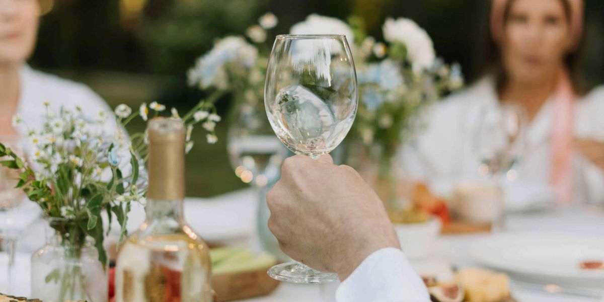 Hand holding a glass of white wine at an outdoor table set with flowers, bread, and cheese.