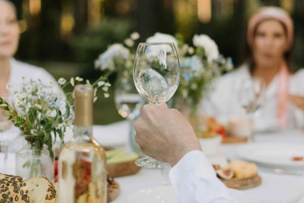 Hand holding a glass of white wine at an outdoor table set with flowers, bread, and cheese.