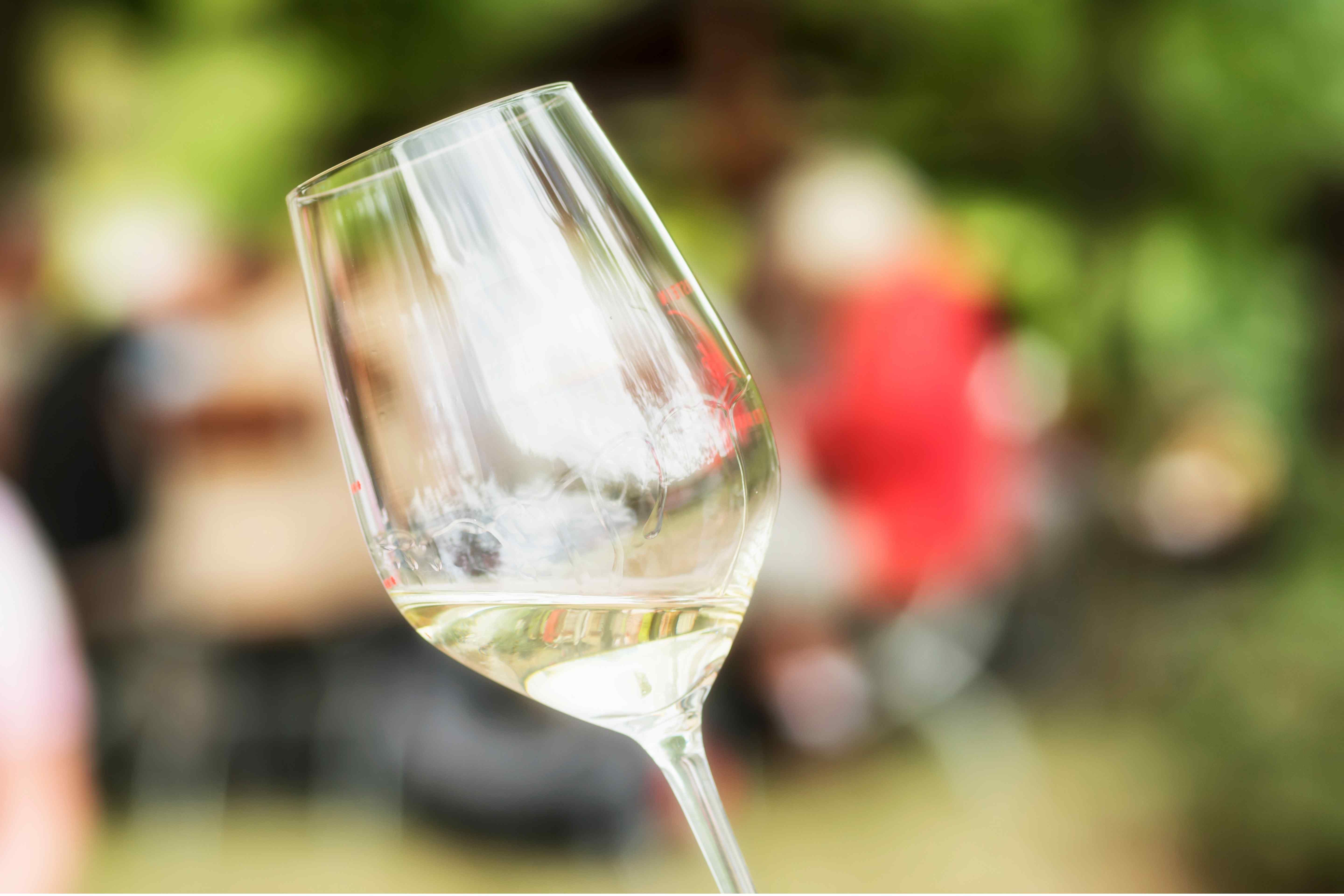 Tilted glass of white wine held outdoors with blurred people in the background.