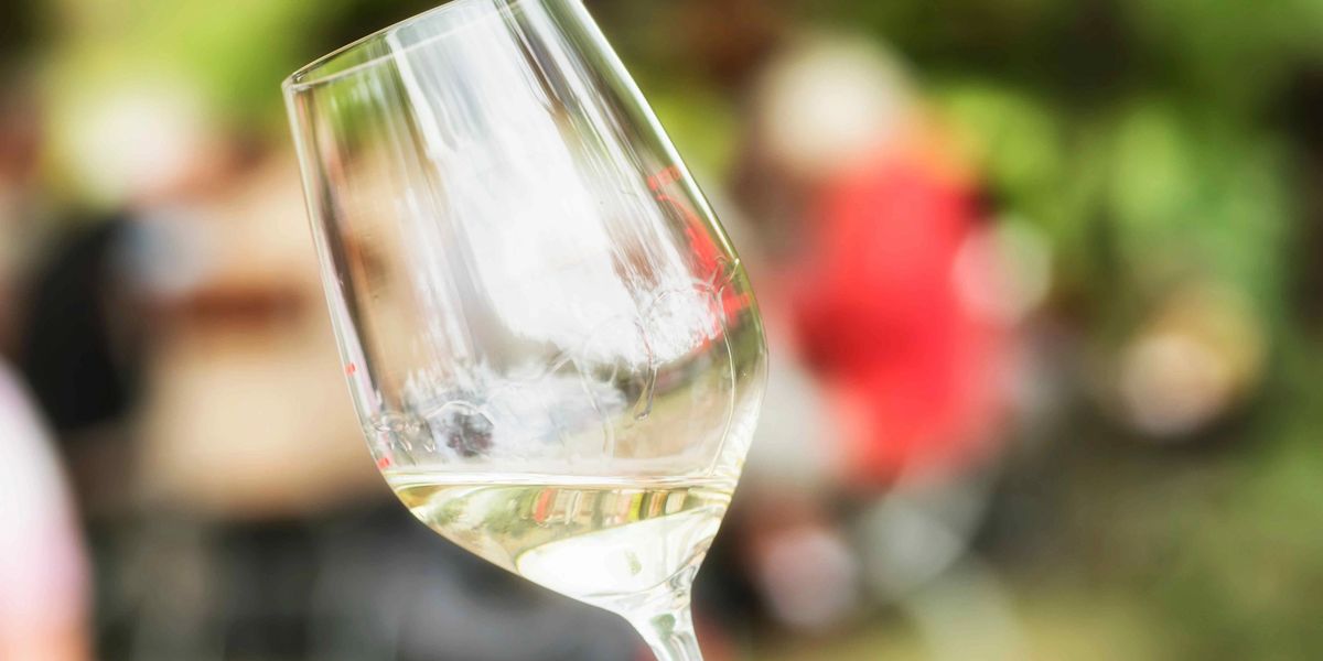 Tilted glass of white wine held outdoors with blurred people in the background.