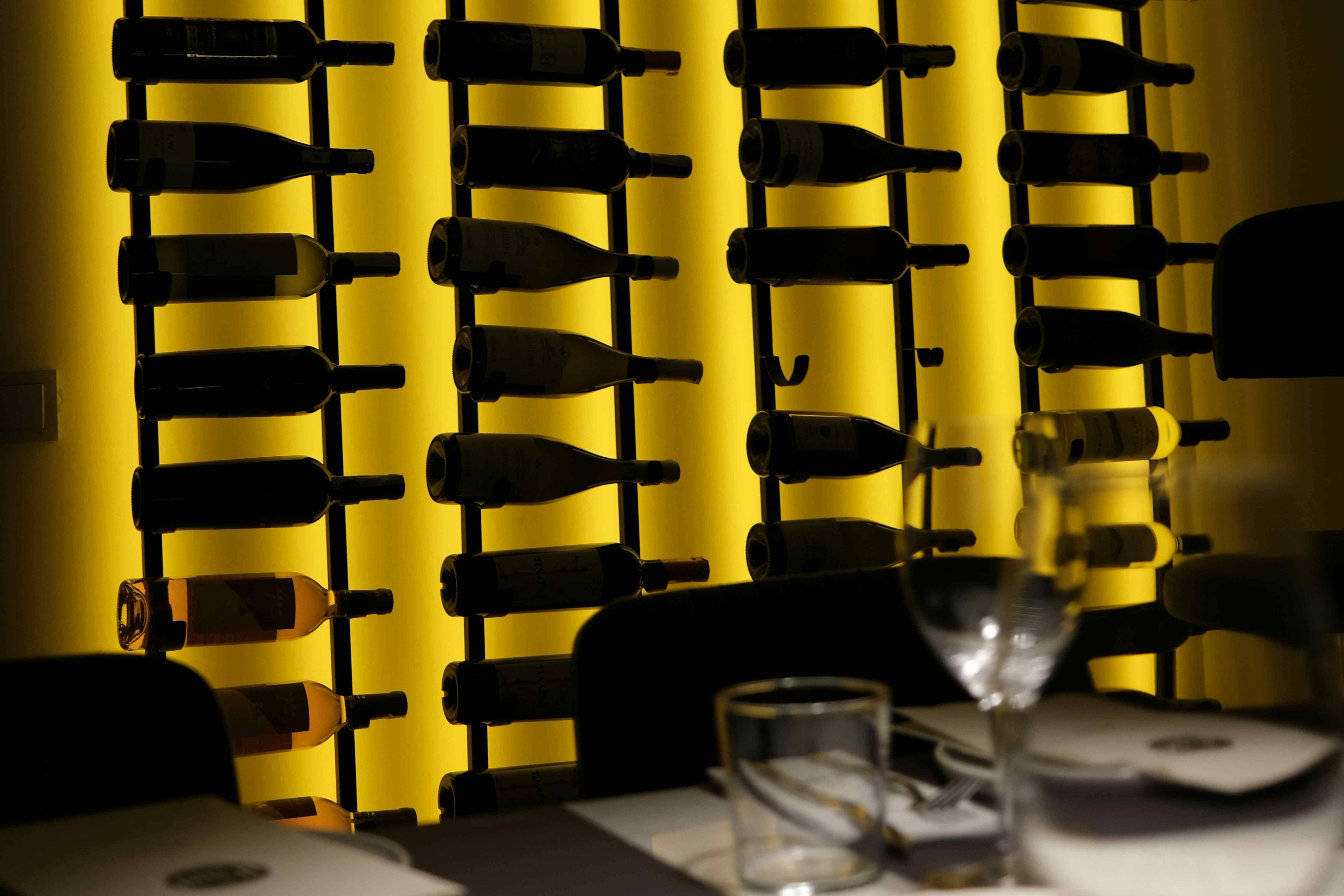 Wine bottles on black metal wall racks against a backlit yellow panel, with a set restaurant table in the foreground.