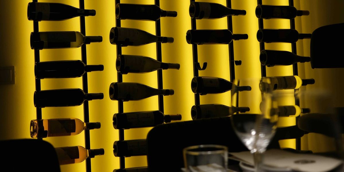 Wine bottles on black metal wall racks against a backlit yellow panel, with a set restaurant table in the foreground.
