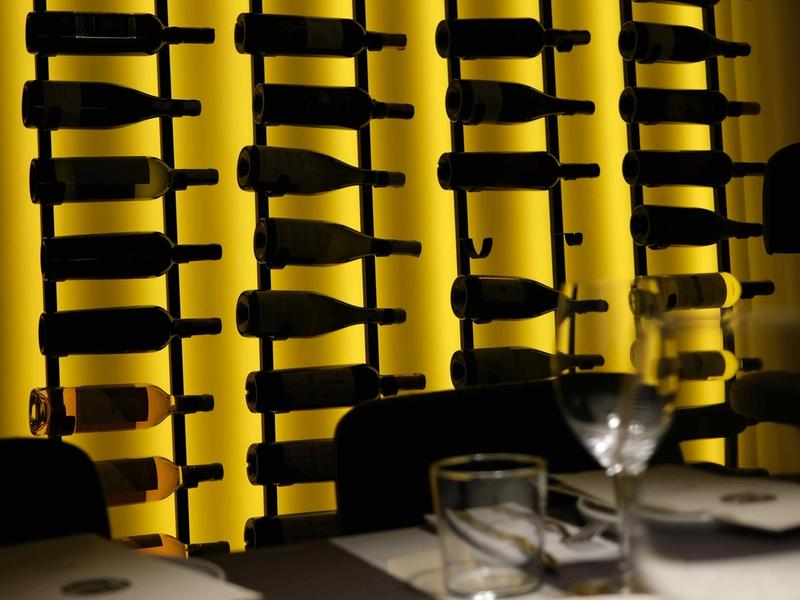 Wine bottles on black metal wall racks against a backlit yellow panel, with a set restaurant table in the foreground.