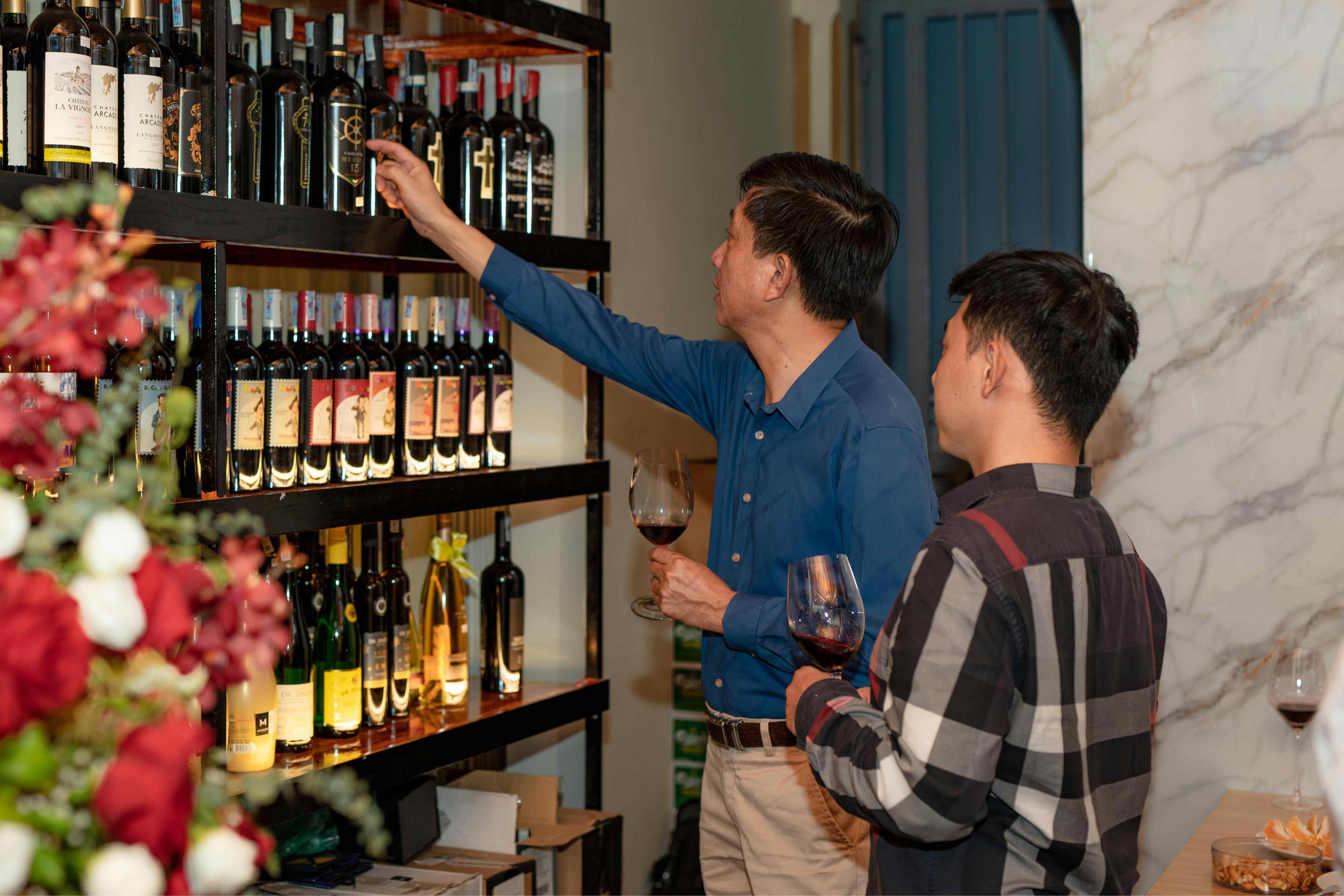 Two men holding glasses of red wine look over bottles on dark shelves, one reaching up to pull a bottle from the top row.