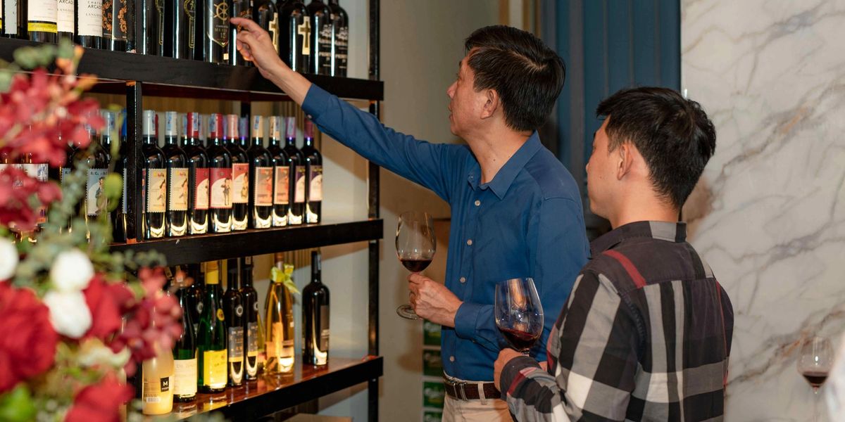 Two men holding glasses of red wine look over bottles on dark shelves, one reaching up to pull a bottle from the top row.