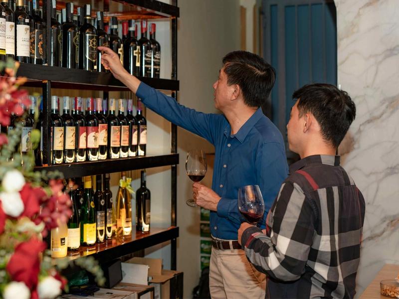 Two men holding glasses of red wine look over bottles on dark shelves, one reaching up to pull a bottle from the top row.