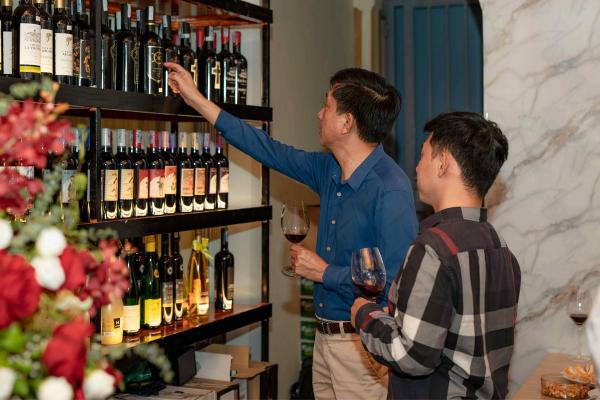 Two men holding glasses of red wine look over bottles on dark shelves, one reaching up to pull a bottle from the top row.