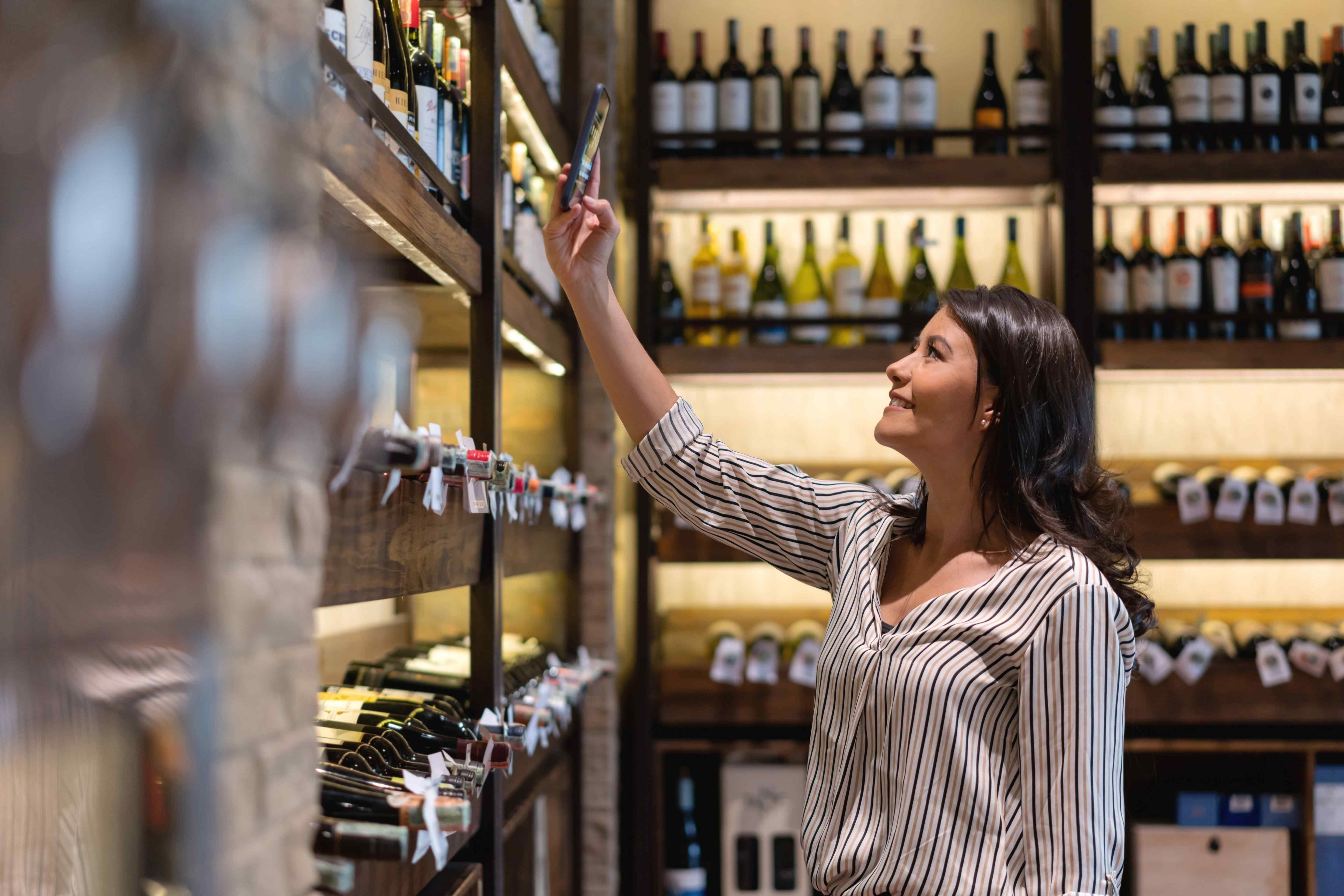 Woman holding up a phone to scan a wine bottle label on a shelf in a wine shop.