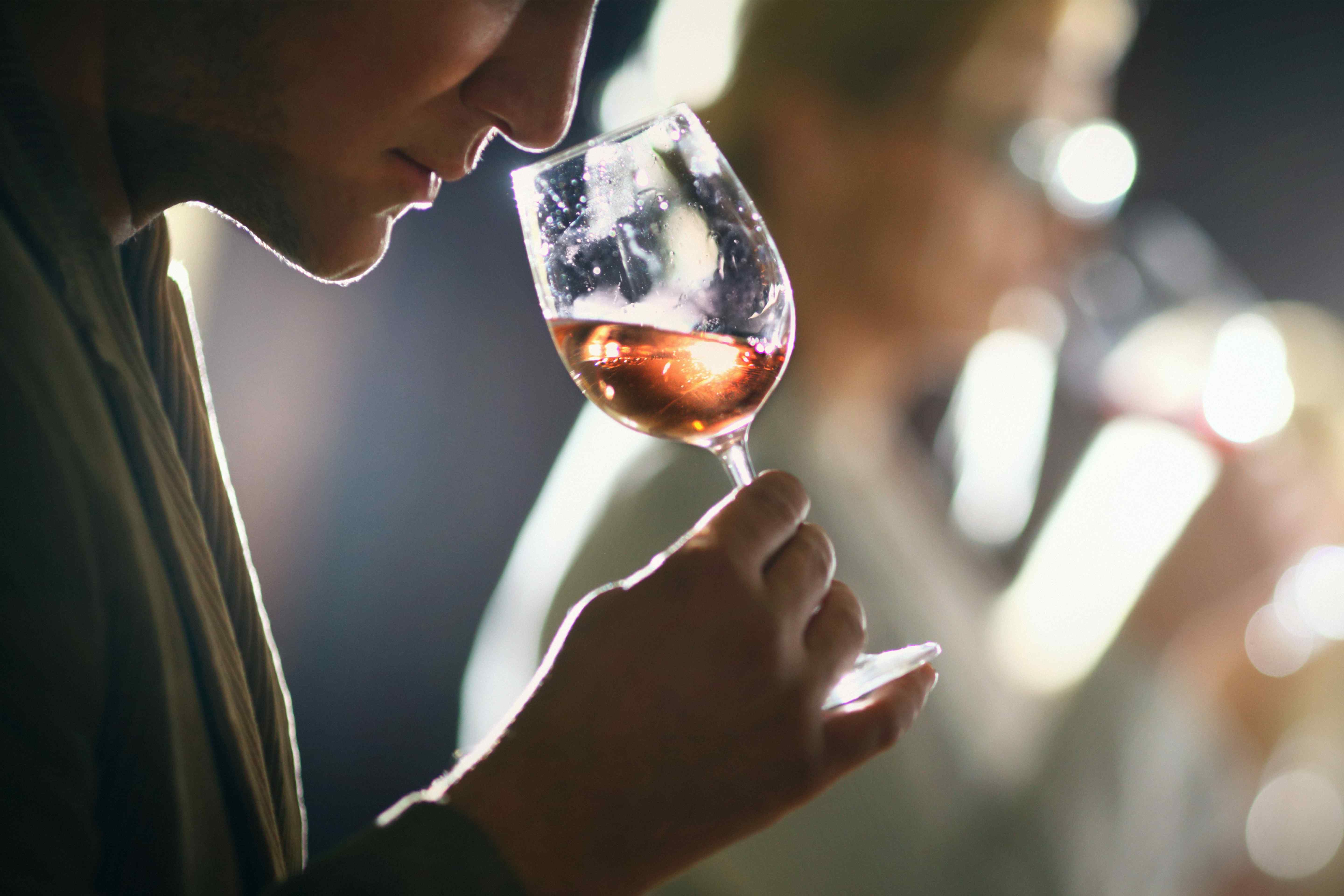 Person smelling a glass of rosé wine at a tasting, backlit with blurred figures in the background.