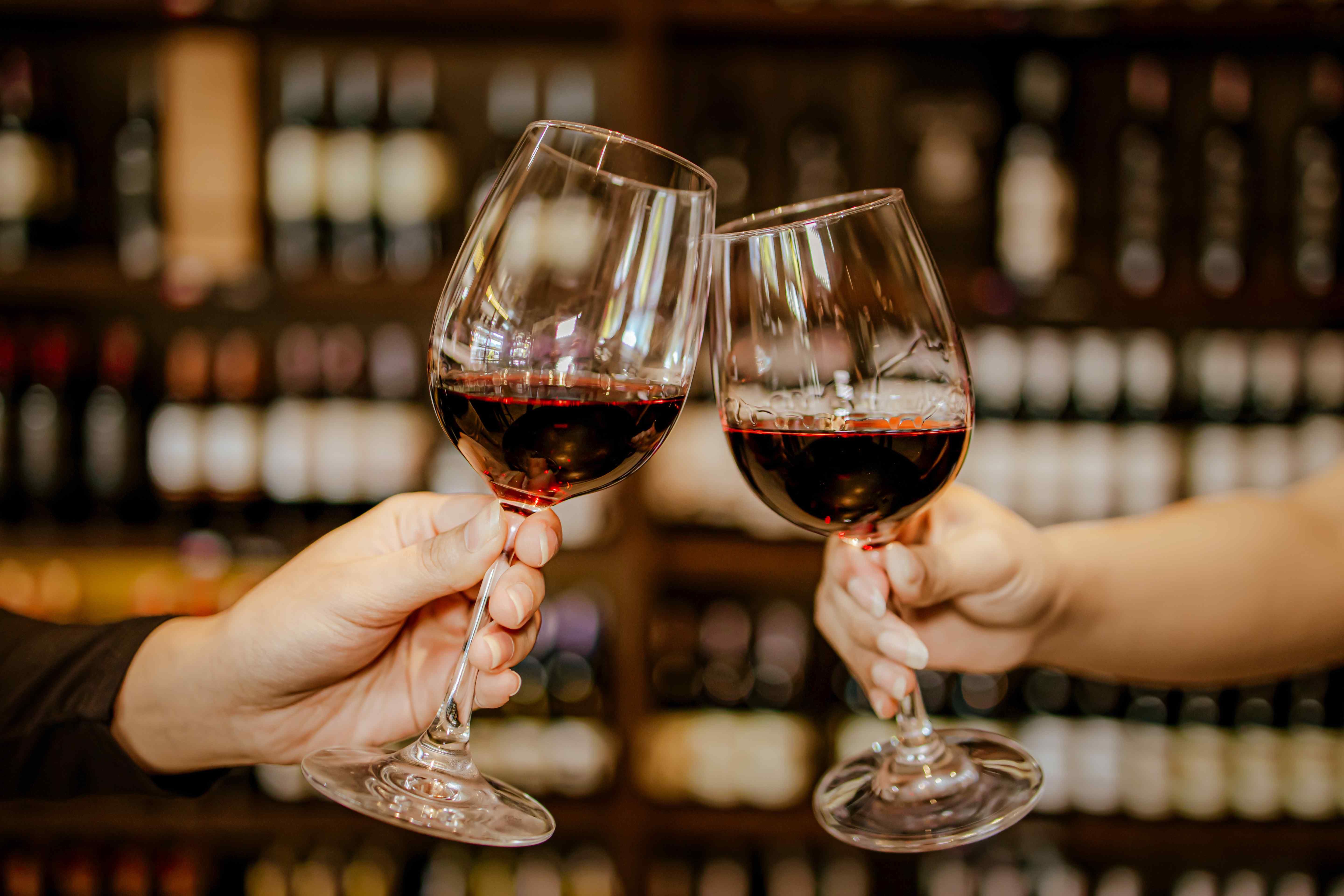 Two hands clink glasses of red wine in front of a blurred wall of wine bottles.