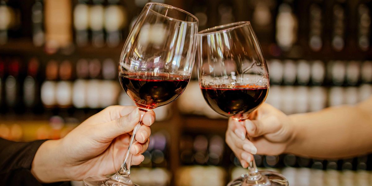Two hands clink glasses of red wine in front of a blurred wall of wine bottles.