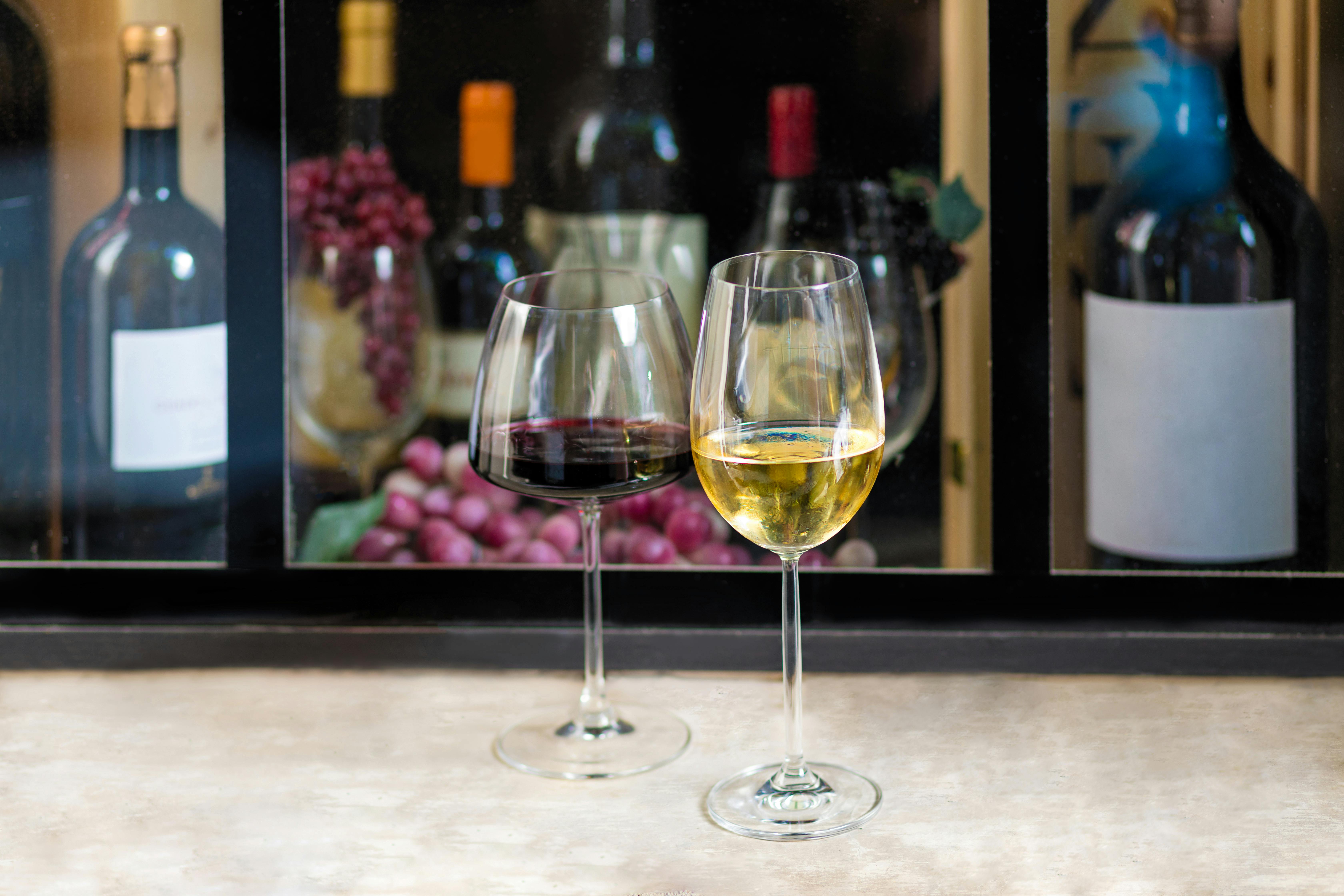 A full glass of red wine and a full glass of white wine rest on a table in front of a wine display shelf.