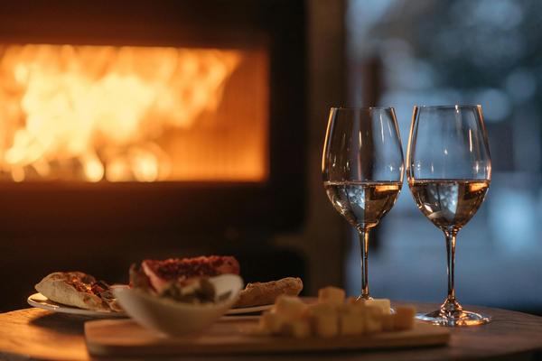 A cozy scene: snacks and full wine glasses sit on top of a coffee table in front of a roaring fireplace.