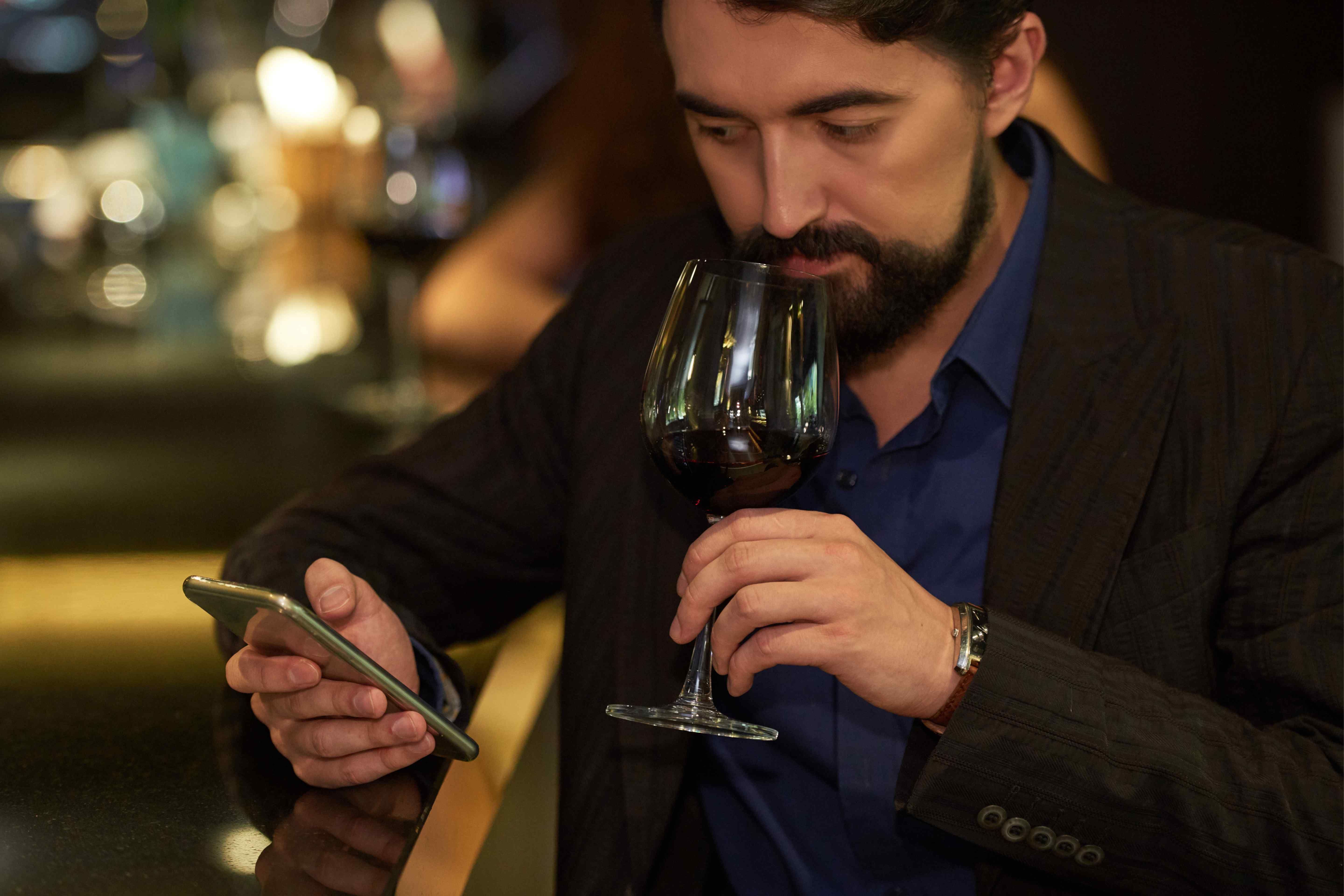 Man checking his phone while smelling a glass of red wine at a dimly lit bar.