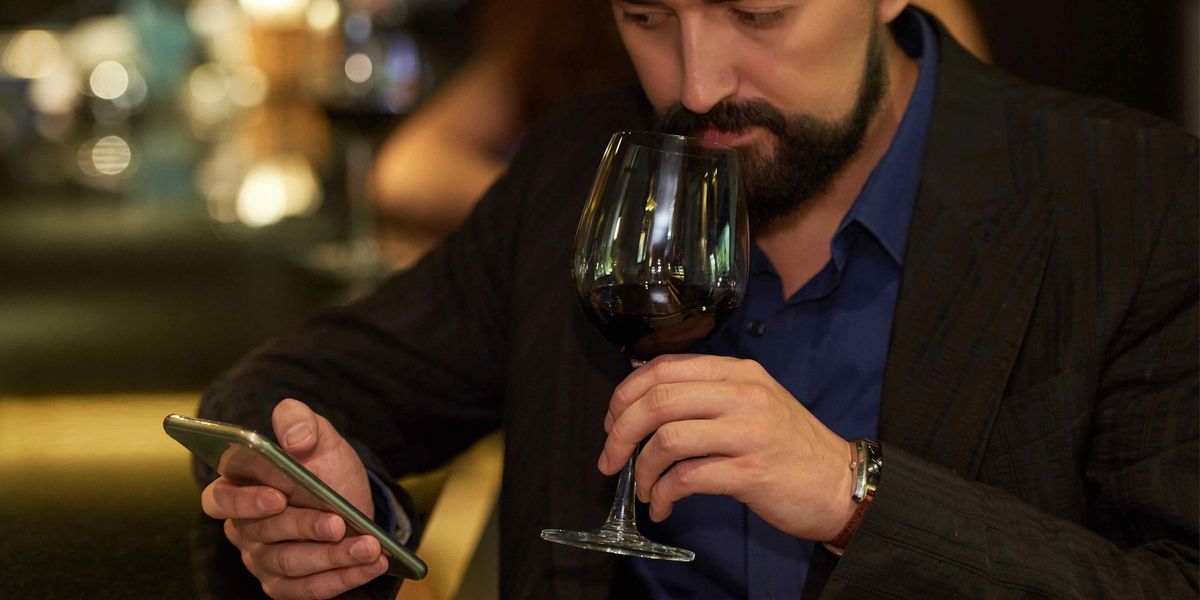 Man checking his phone while smelling a glass of red wine at a dimly lit bar.