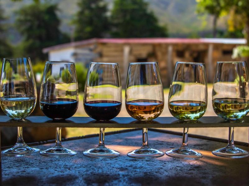 Six wine glasses of varying colors, from white to red, lined up outdoors in evening light.