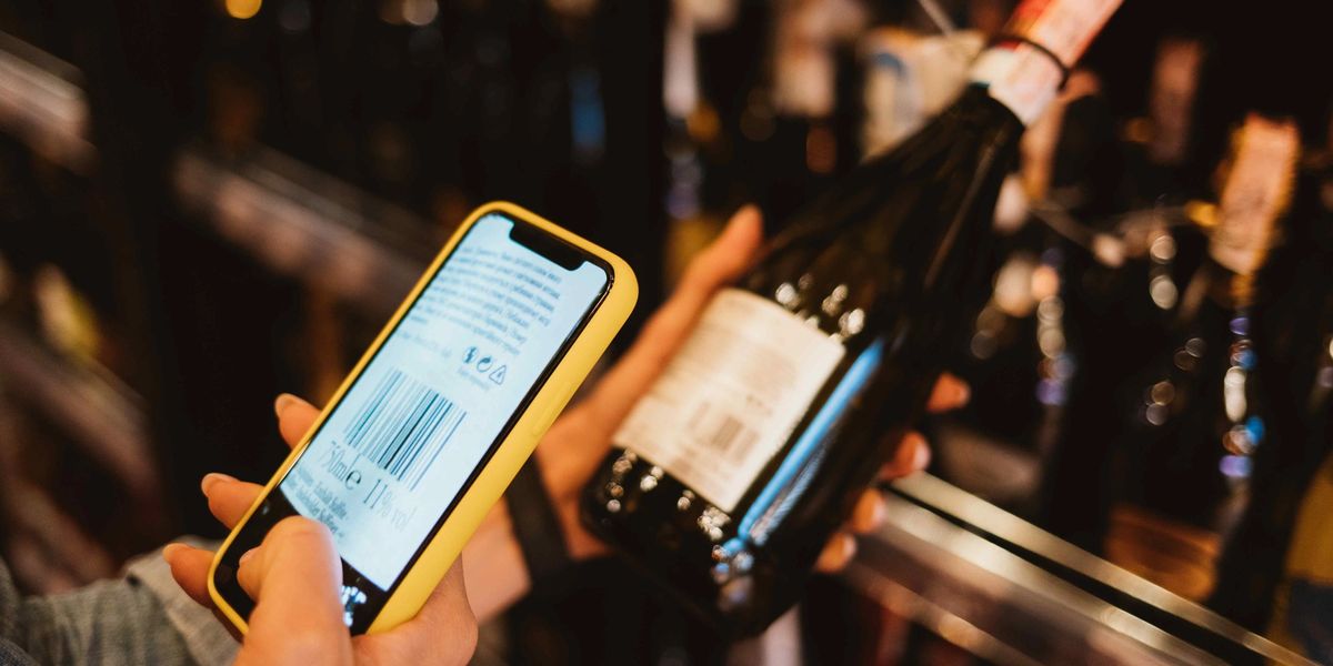 A hand holds a yellow-cased phone up to scan the barcode on a wine bottle’s back label in a store aisle.