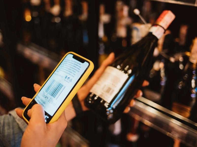 A hand holds a yellow-cased phone up to scan the barcode on a wine bottle’s back label in a store aisle.
