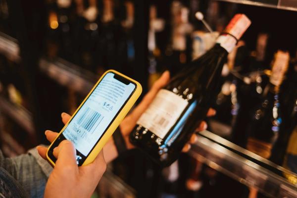 A hand holds a yellow-cased phone up to scan the barcode on a wine bottle’s back label in a store aisle.