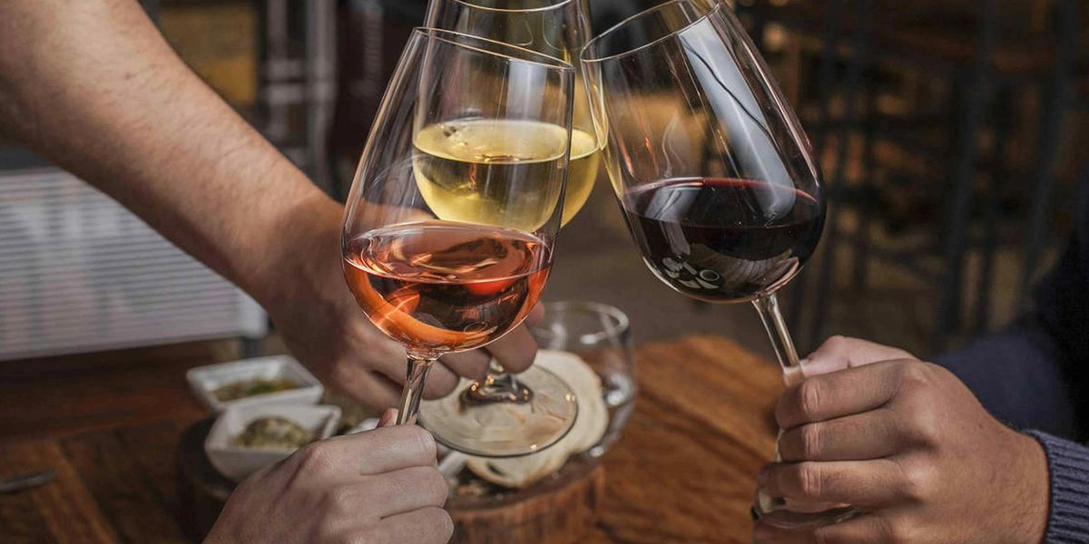 Three hands clinking glasses of rosé, white and red wine over a wooden restaurant table with small plates.