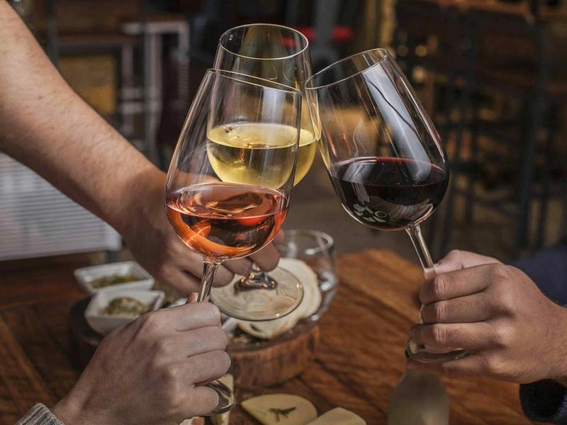 Three hands clinking glasses of rosé, white and red wine over a wooden restaurant table with small plates.