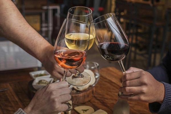 Three hands clinking glasses of rosé, white and red wine over a wooden restaurant table with small plates.